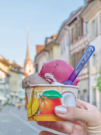 Eine Person hält einen Glacebecher mit 3 Kugeln in eine Gasse.