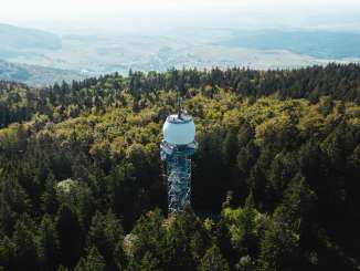 Aus einem Wald ragt ein hoher Aussichtsturm aus Metall. Über der Aussichtsplattform ist eine grosse weisse Kugel.