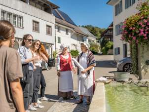 Wöschwiiber-Führung Wilchingen An einem Dorfbrunnen stehen zwei Frauen verkleidet als Wöschwiiber mit Gewand und Haube. Eine Gruppe hört ihnen beim Geschichten erzählen zu