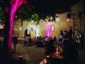 Festbänke in einem beleuchteten Innenhof. Die Bänke sind gefüllt mit Menschen. Es ist eine kleine Bühne aufgestellt. Darauf musiziert ein Musiker.