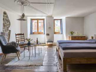 Baderhaus Neunkirch, Ferien im Baudenkmal In einem Zimmer mit Holzboden steht ein Doppelbett, ein Sessel und ein Holzstuhl. In der weiss verputzen Wand hat es ein Loch, wo die historische Mauer freigelegt ist.