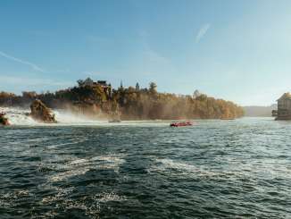 Blick auf den grössten Wasserfall Europas. Im Becken fahren Boote, die Bäume tragen goldiges Herbstlaub und man sieht das Schloss Laufen und Schlössli Wörth.
