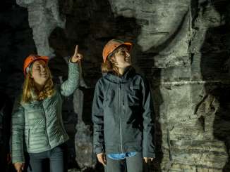 Drei Personen stehen in einem Bergstollen. Alle tragen einen Helm. Jemand zeigt mit dem Zeigefinger an die Decke.