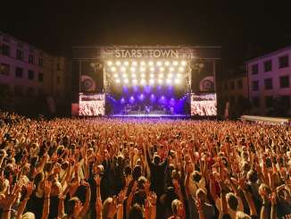 Stars in der Stadt Viele Personen mit den Armen in der Luft sind an einem Konzert mit einer Band auf einer grossen Bühne.
