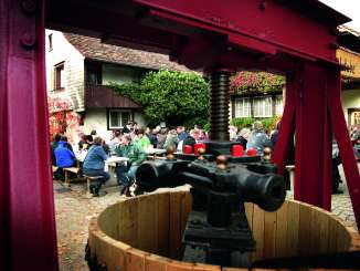 Ein Fest auf einem Vorplatz. Eine grosse Fruchtpresse ist im Vordergrund zu sehen.