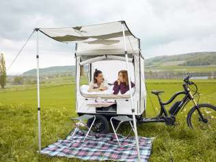 Zwei Freundinnen liegen in einem Zeltaufbau auf einem Cargovelo. Davor ist eine Picknick-Decke mit Campingstühlen ausgebreitet.