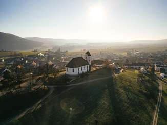 Auf einem Hügel mit Reben stehe eine Kirche. Dahinter sieht man das Dorf.