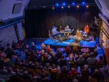 Eine kleine Bühne mit vier Musiker. Der volle Saal hört gespannt zu. Die Beleuchtung ist blau und rot.