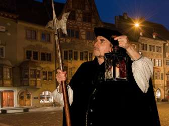 Eine Person in einem schwarzen Umhang gekleidet steht in der Dämmerung in der Altstadt. In der einen Hand trägt sie eine Laterne in der anderen eine Lanze.