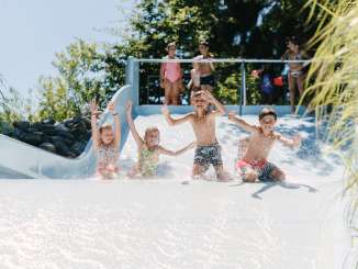 Vier Kinder rutschen in einem Freibad gemeinsam eine breite Rutschbahn hinunter.
