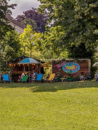 In einem Park steht ein Holzhäusschen und ein Anhänger, die als Pop-Up Café dienen. Davor stehen viele bunte Liegestühle und Holztische mit Stühlen.