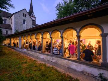 Diverse Stände, wo man Wein degustieren kann, sind im Kreuzgang zu Allerheiligen aufgestellt. Personen stehen an den Stände und trinken Wein.