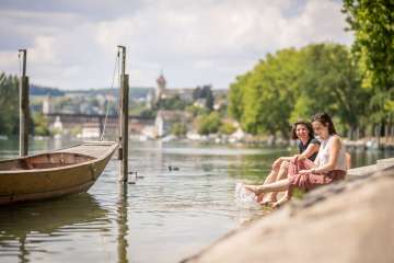 Zwei Frauen sitzen am Ufer eines Flusses und halten ihre Füsse zur Abkühlung ins Wasser. In der Ferne ist eine Altstadt zu sehen.