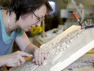 Eine Frau sitz vor einer Holplatte. Mit verschiedenen Werkzeugen schnitzt sie Muster.