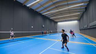 Eine Halle mit vier blauen Badminton-Felder. Darauf spiel Personen Badminton.