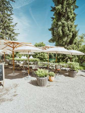 Holzstühle sowie Tische sind gedeckt und weisse Sonnenschirme spenden Schatten. Die Restaurantsterrasse liegt direkt am Rhein mit viel Grün umgeben.