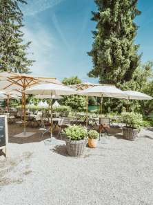 Holzstühle sowie Tische sind gedeckt und weisse Sonnenschirme spenden Schatten. Die Restaurantsterrasse liegt direkt am Rhein mit viel Grün umgeben.
