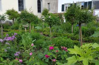 Ein Garten mit blühenden Blumen in Violett und Pink. Im Hintergrund steht ein altes Haus.