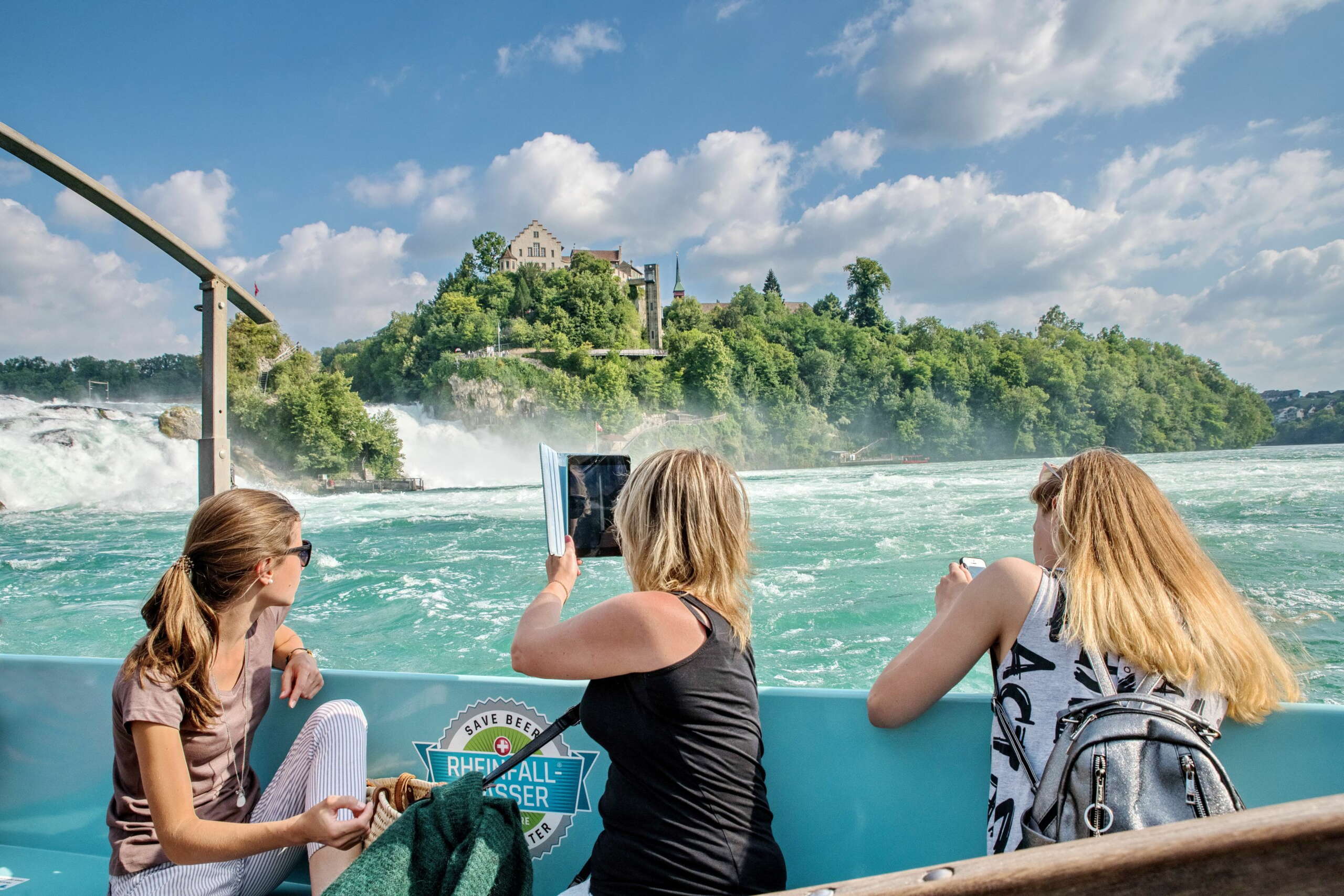Drei Touristinnen machen aus einem Boot Fotos vom Rheinfall. Im Hintergrund ist das Schloss Laufen.