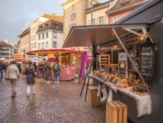 Auf einem Platz stehen viele Marktstände. Personen laufen vorbei.