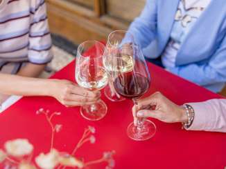 Drei Personen stossen mit zwei Gläser Weisswein und einem Glas Rotwein an. Sie sitzen an einem kleinen roten Tisch.