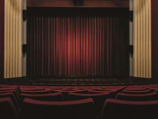 Ein leerer Saal mit Bühne. Alle Stühle sind rot gepolstert. Der zugezogene Vorhang ist ebenfalls rot.