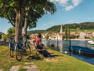 Zwei Freudinnen sitzen auf einer Holzbank am Rhein mit Blick auf Stein am Rhein. Hinter ihnen stehen zwei E-Bikes.