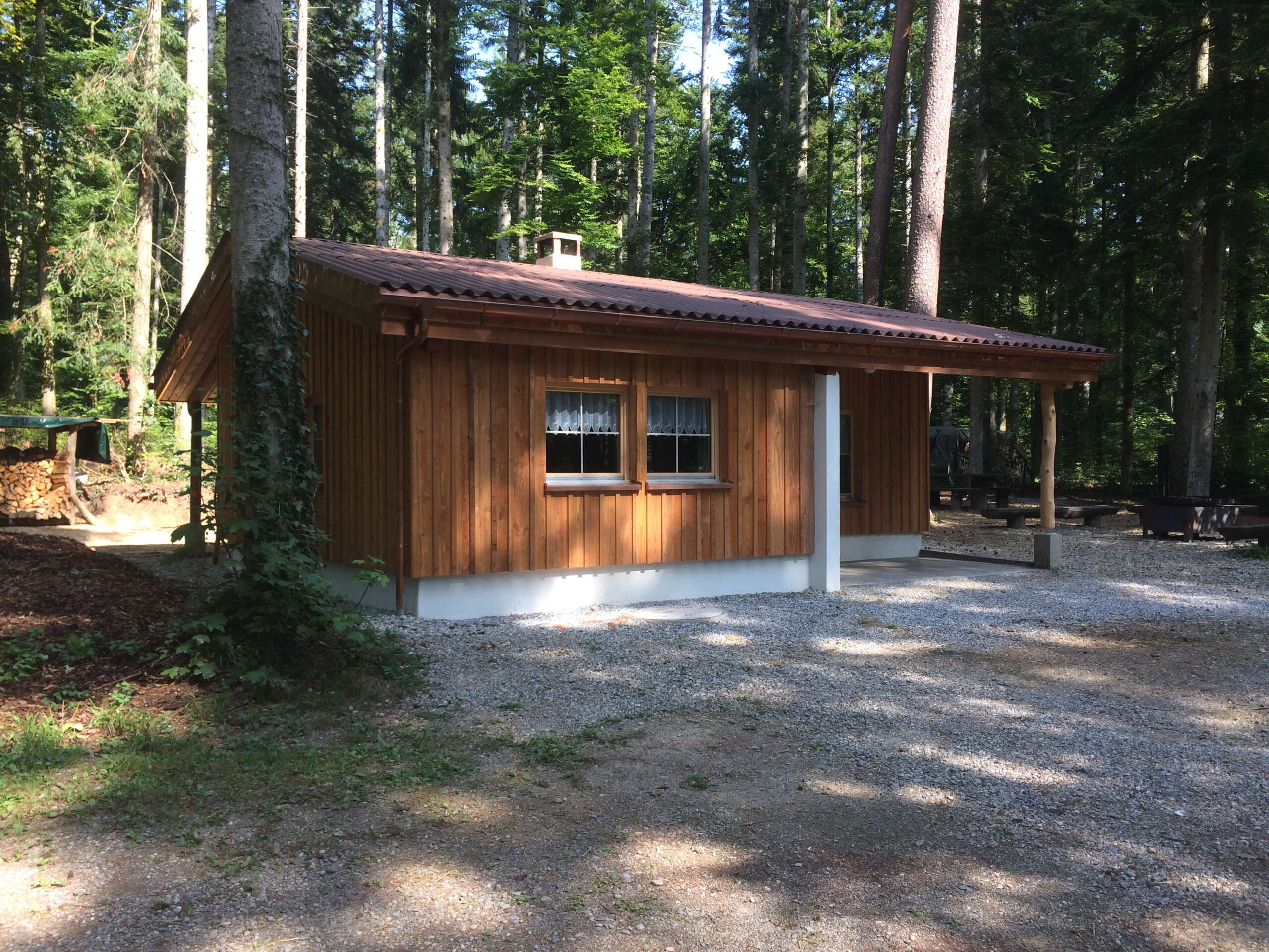 Kleine Waldhütte mit einer Holzverkleidung und einem Vordach/Unterstand. Rundherum hat es Wald.