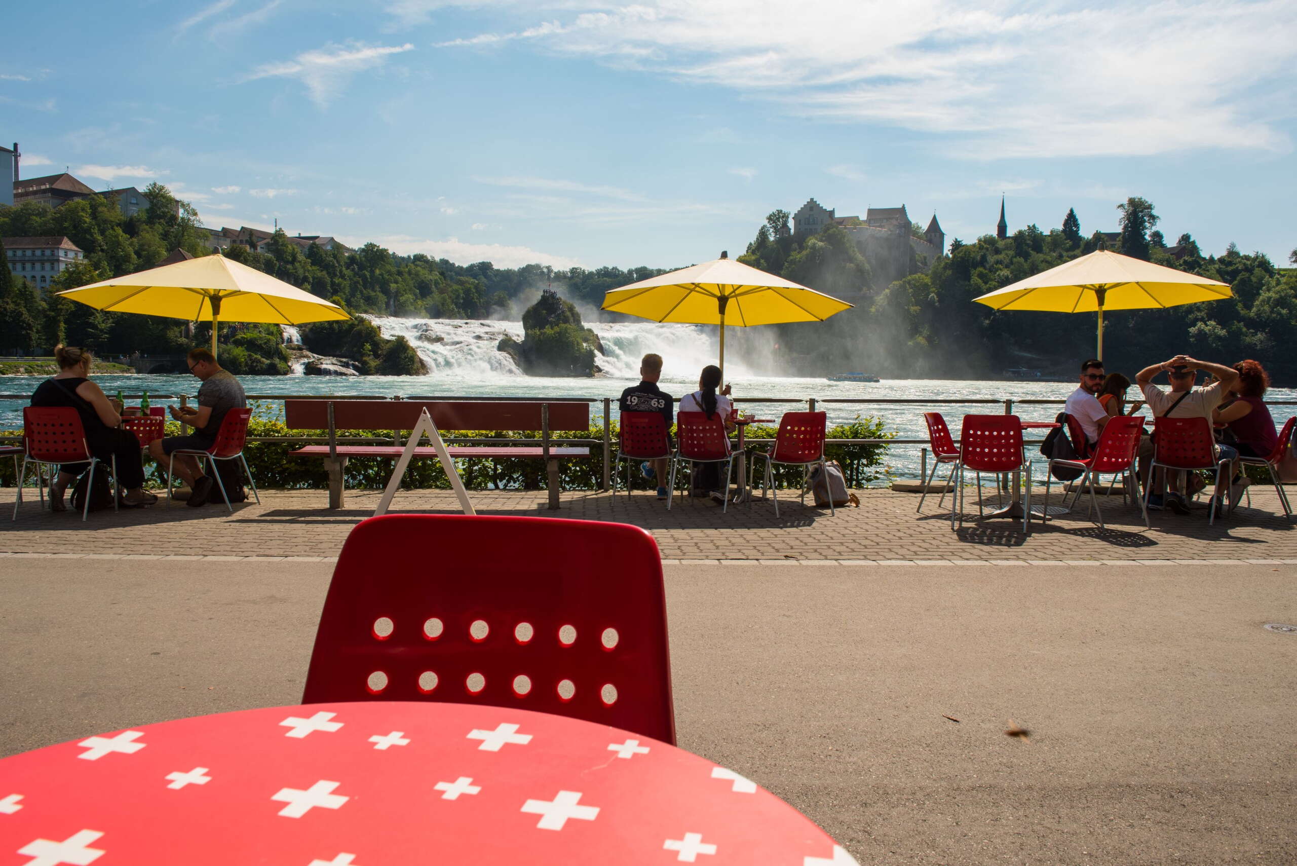 Rote Tische mit weissen Schweizer Kreuzen, rote Stühle und gelbe Sonnenschirme vom Restaurant Park am Rheinfall. Im Hintergrund ist der Rheinfall.