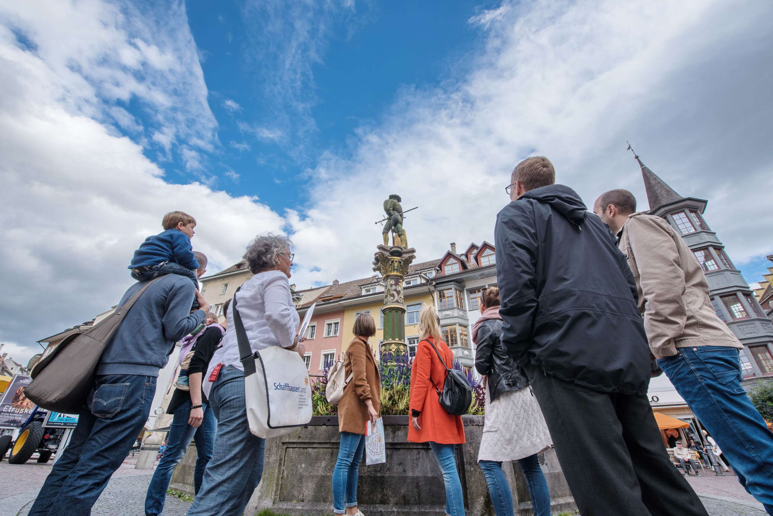Stadtführung durch die Schaffhauser Altstadt. Die Gruppe und der Guide stehen auf dem Fronwagplatz vor dem Landsknechtbrunnen und betrachten die Landsknechtfigur.
