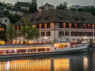 Blick vom Rhein bei Dämmerung auf ein beleuchtetes Schiff und ein Riegelhaus direkt am Rhein.