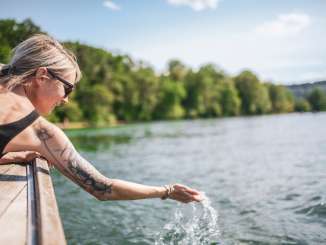 Eine Frau trägt einen schwarzen Badeanzug und Sonnenbrille. Sie liegt auf einem Holzboot und hält eine Hand ins Wasser.