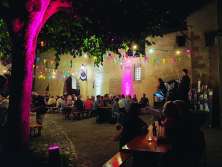 Festbänke in einem beleuchteten Innenhof. Die Bänke sind gefüllt mit Menschen. Es ist eine kleine Bühne aufgestellt. Darauf musiziert ein Musiker.