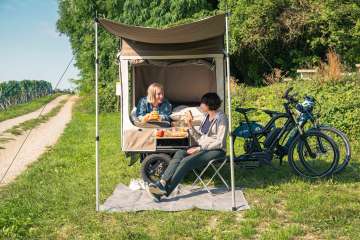 Velosafari Schaffhauserland Auf einer Wiese neben Reben ist ein Cargovelo mit aufgeklapptem Zeltaufbau. Eine Peron liegt im Bett und sitz auf einem Campingstuhl davor.