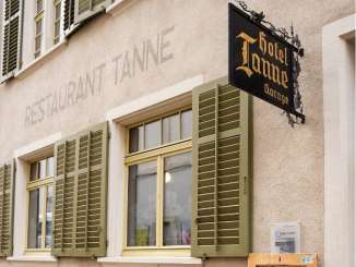 Auf einer Hausfassade steht gross "Restaurant Tanne". Es hat grüne Fensterläden und ein eisernes schwarzes Schild mit goldiger Schrift "Hotel Tanne".