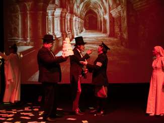 Sechs verkleidete Theaterspielende. Ein grossen Bild von einem Kreuzgang im Hintergrund. Eine Nonne, ein Polizist, ein Herr mit Zilinder, ein Herr mit einer Torte in der Hand.