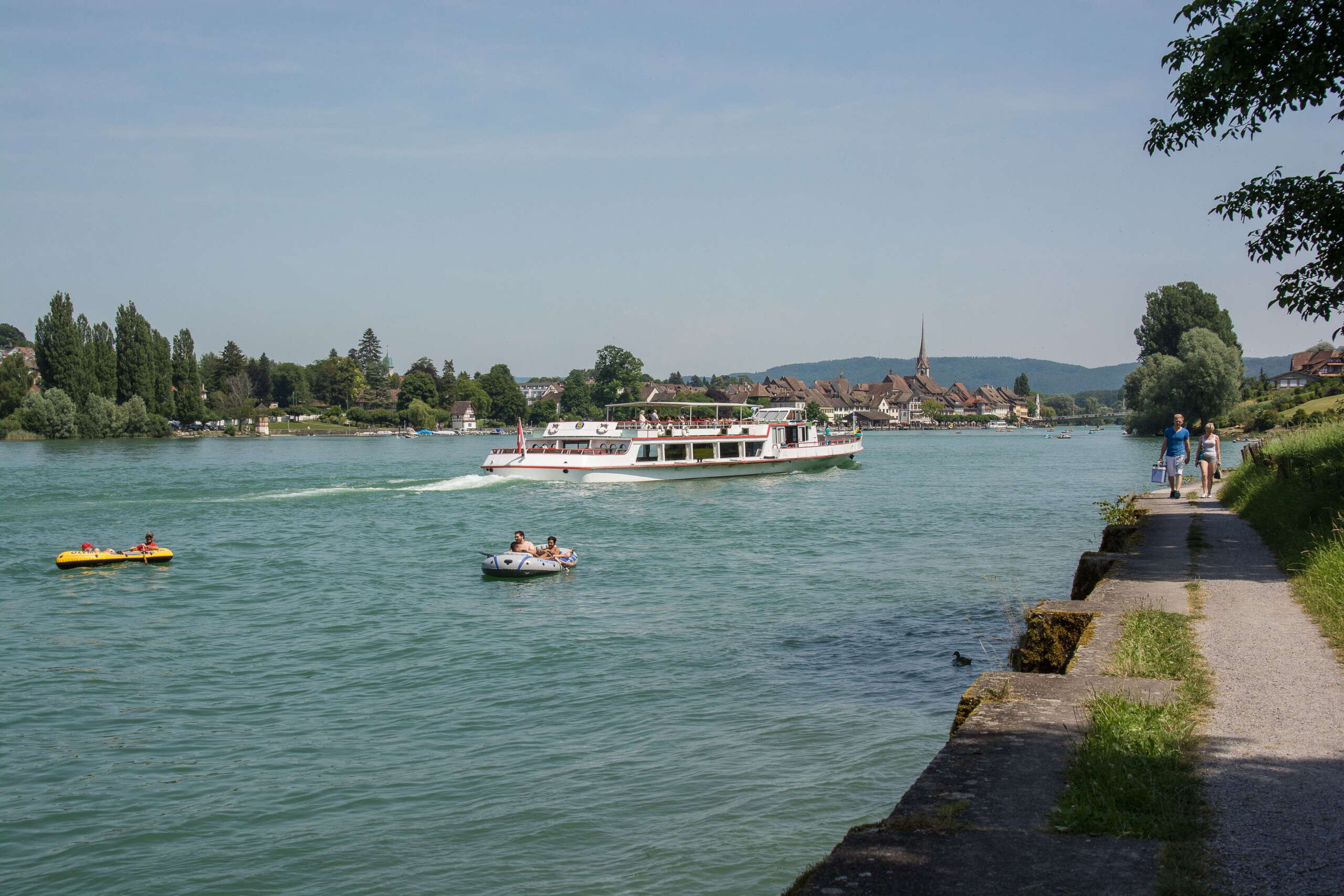 Kursschiff der URh fährt Richtung Stein am Rhein. Zwei Gummiboote treiben rheinabwärts.