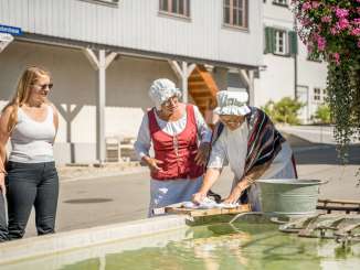 Zwei verkleidete Frauen welche Wäsche mit einem Waschbrett am Brunnen waschen und miteinander sprechen. Die anderen Personen hören gespannt zu.