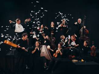 Mehrere schwarz gekleidete Musizierende sind vor einem schwarzen Hintergrund, haben Instrumente in der Hand und weisse Federn fliegen in der Luft.