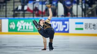 Lukas Britschgi während seinem Eiskunstlaufprogramm. Es trägt eine schwarze Hose und ein schwarzes Oberteil ohne Ärmel mit goldigen Verziehrungen.