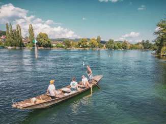 Weidlingsfahrt auf dem Rhein Drei Personen und ein Hund sitzen im Weidling. Das Holzboot treibt gemütlich den Rhein hinunter. Eine Frau steht, hat ein Ruder in der Hand und steuert das Boot.