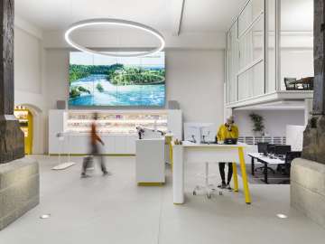 Visitor Centre Schaffhausen Das ist der Schalterbereich des Visitor Centres Schaffhausen. An einem Stehtisch steht eine Frau in senfgelben Pullover und telefoniert. Eine weitere Person läuft zum freien Schalter.