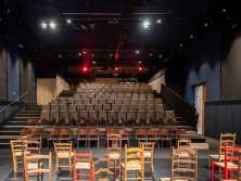 Eine Bühne mit vielen verschiedenen Holzstühlen. Eine bestuhlte Tribüne. Der Eingang befindet sich rechts.
