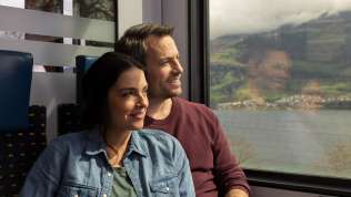 Ein Mann und eine Frau sitzen nebeneinander im Zug und schauen aus dem Fenster auf die Landschaft mit Hügel, Dörfer und einem See.