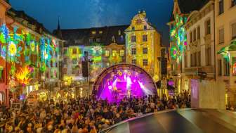 Stars in der Stadt Ein Platz zwischen Häusern. Die Fassaden sind farbig beleuchtet. In der Mitte ist eine Bühne mit Musiker-/innen. Der Platz ist gefüllt mit Menschen.