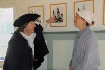 Eine Person mit schwarzem Umhang und schwarzem Hut zeigt auf ein Bild mit einem Turm der brennt darauf. Eine zweite Person mit einem Hellen Kleid und weissem Kopftuch hört gespannt zu.