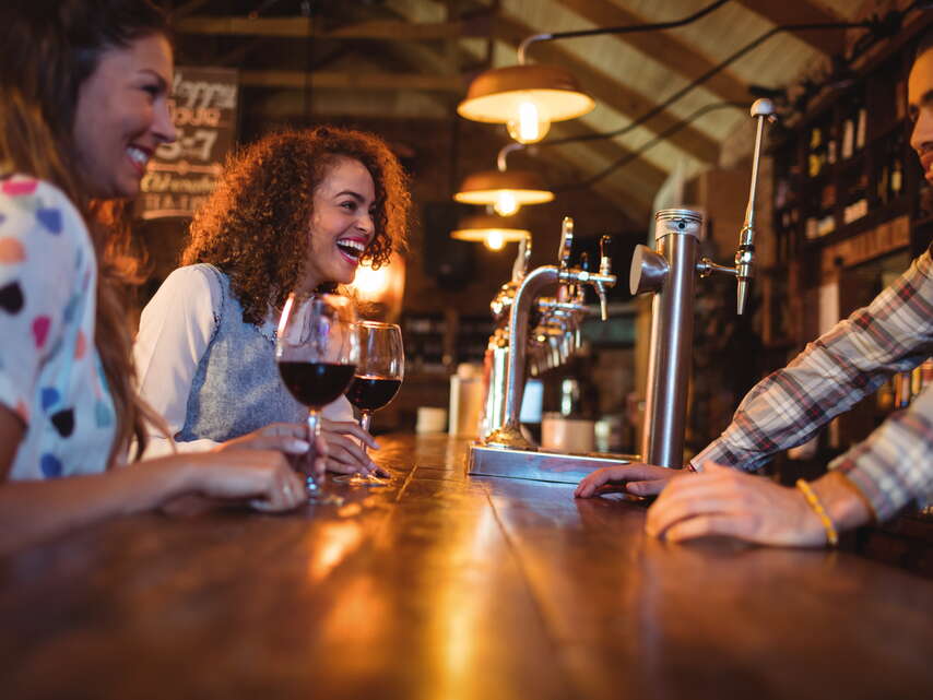 Zwei Freundinnen geniessen ein Glas Wein in einer Bar und sich fröhlich am Lachen und unterhalten sich mit dem Barkeeper.