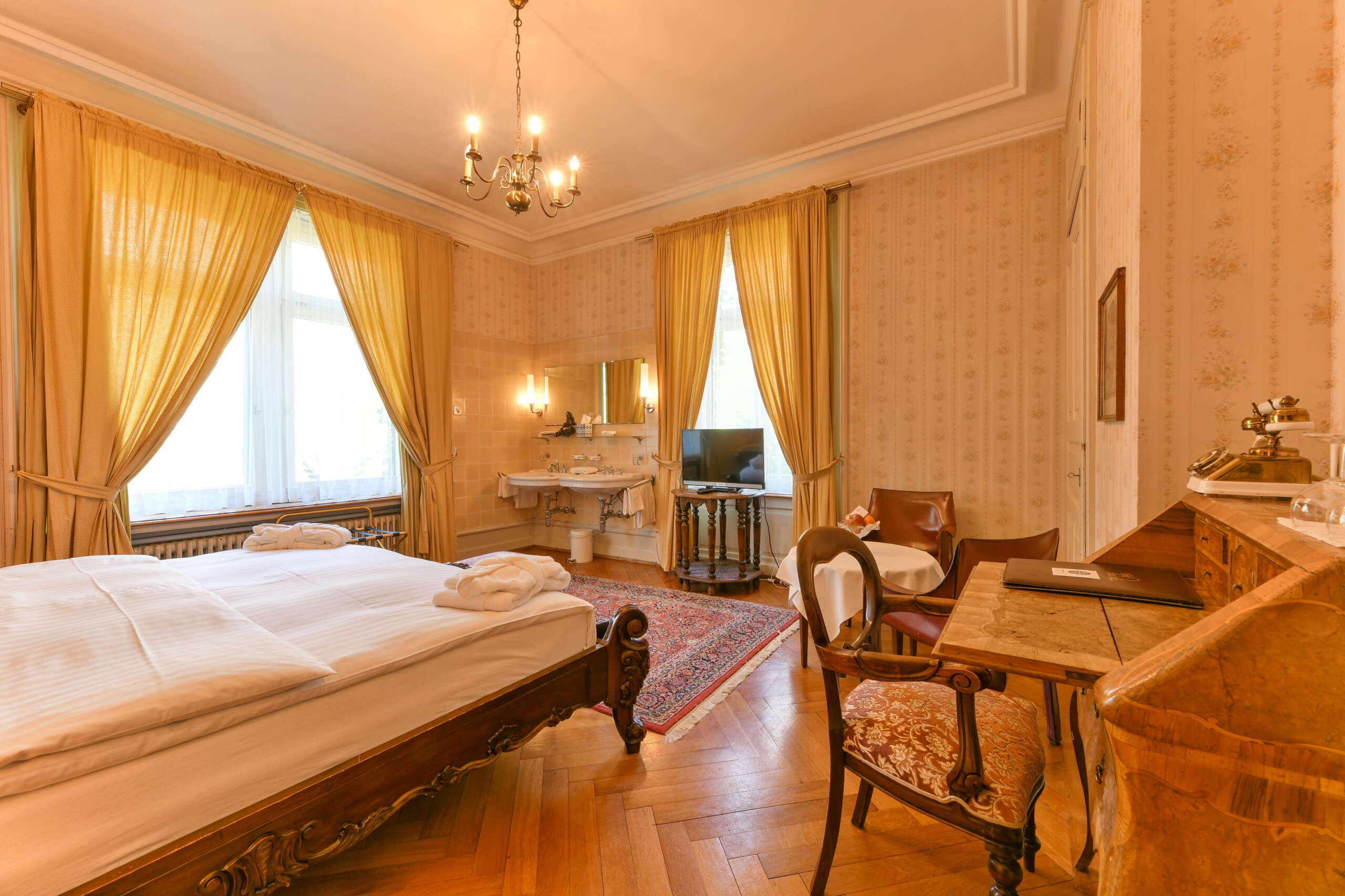 Ein Hotelzimmer stilvoll eingerichtet mit Biedermeiermöbeln, gelben Vorhängen, einem Teppich und passender Tapete.