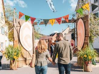 Ein Paar spaziert Händchenhaltend auf ein Tor zu. Über dem Tor hängt der Schriftzug: "Herzlich Willkommen". Das Tor ist mit Blumen geschmückt.