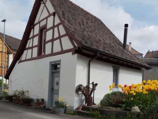 Schlafen im Wöschhüsli Ein altes kleines Haus mit weisser Fassade und braunen Balken. Rechts davon ist ein Garten mit diversen Blumen.